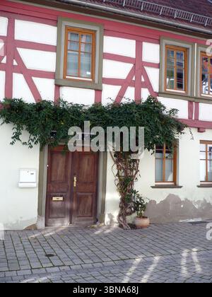 Idstein, Germany, A charming half-timbered house with a red and white exterior. The house features a wooden door with a gold-colored handle and a small window above it. A large green vine with small yellow flowers is growing on the side of the house, partially covering the door. A small, light-colored mailbox is attached to the wall next to the door. The house is on a cobblestone street, with a small, light-colored planter with a plant in a terracotta pot placed in front of the house. The house has multiple windows with white frames and brown or dark brown wooden frames. The roof is dark Stock Photo