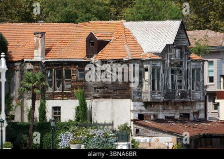 Istanbul, Turkey. The largest city in Turkey and one of the world’s few transcontinental cities, spanning both Europe and Asia across the Bosphorus Strait., A dilapidated house with a red tile roof and a white metal roof. The house is in a state of disrepair, with a large section of the front wall missing, revealing the underlying structure. The house is surrounded by lush greenery, including trees and bushes, and a portion of a white building is visible to the right. A palm tree is also visible in the foreground. The house is partially obscured by a dark-colored fence or railing. The house's Stock Photo