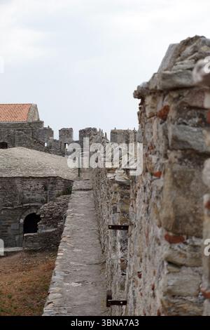 Kavala, Greece, A stone wall, constructed from various shades of gray and brown stones, with a rough texture. The wall is flanked by two structures, one with a red tile roof and the other with a green roof. A stone pathway, made of uneven stones, runs along the wall, leading towards the structures. The sky is overcast, casting a soft light over the scene. The perspective of the image is from the side of the wall, looking towards the structures and the sky. An artist's eye & imagination shaped Stock Photo