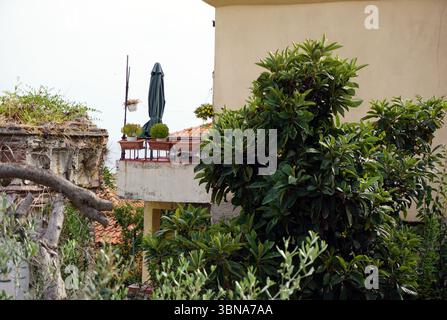 Kavala, Greece, A balcony with a green umbrella and a few potted plants, including a small tree or shrub, on a beige or cream-colored building. The balcony is part of a larger building with a cream or beige facade. The balcony is elevated above lush greenery, including a small tree or shrub in a pot. The building has a terracotta-colored roof. The sky is overcast, casting a soft light over the scene. An artist's eye & imagination shaped Stock Photo