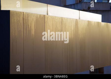 Cyprus, Larnaca, The image shows a section of a building with a series of rectangular panels, likely made of metal or a similar material. The panels are arranged in a row and have a light beige or tan color. The panels are slightly offset from each other, creating a sense of depth. The panels are adorned with a textured pattern that resembles dripping water, adding a unique visual element to the structure. The exterior is primarily a light beige or tan color, with a dark gray or black section on the left side. The background is out of focus, but it appears to be a building with a Stock Photo