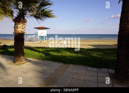 Cyprus, Larnaca, A serene beach scene. In the foreground, a white lifeguard tower with a red roof stands on a sandy beach, with a metal railing around its base. A small white object, possibly a trash can, is located near the tower. Two palm trees frame the scene, with their fronds reaching towards the clear blue sky. The ocean stretches out in the background, its surface calm and inviting. The beach is bordered by a paved walkway made of light-colored stone or concrete pavers. The grassy area in the foreground is a vibrant green, contrasting with the sandy beach and the clear blue Stock Photo