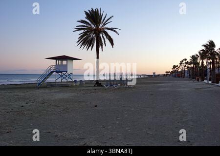 Cyprus, Larnaca, A serene beach scene at sunset. The sky is painted in hues of orange and pink, casting a warm glow over the tranquil setting. The beach is lined with palm trees, their silhouettes standing tall against the sky. A lifeguard tower, painted in white with a red roof, is situated on the left side of the image. A set of stairs leads up to the tower. A row of beach chairs is neatly arranged in the middle ground, and a line of beach umbrellas is visible in the background. The beach is mostly empty, with a few people visible in the distance. The ocean stretches out to the horizon, its Stock Photo