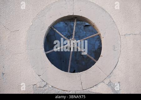 Cyprus, Larnaca, A round window set into a light beige or cream-colored wall. The window is framed by a metal grate with a circular design, and the window opening reveals a dark blue or black interior. The wall surrounding the window is made of concrete or a similar material and shows signs of age and weathering, with cracks and discoloration. The window is centrally located within the image, drawing the viewer's attention to its details. An artist's eye & imagination shaped caption. Larnaca, Cyprus, a coastal gem rich in history, beach life, and cultural charm. One of Cyprus’s oldest cities, Stock Photo