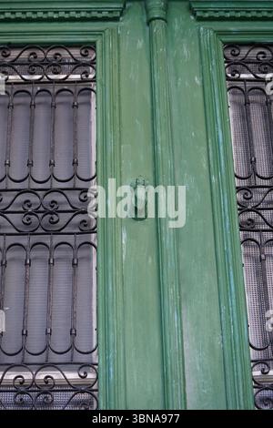 Cyprus, Larnaca, A close-up view of a green door with a black metal door knocker. The door knocker is a small, hand-shaped object, possibly made of wood or metal, and is affixed to the door. The door is framed by a black metal grate with a decorative, swirling pattern. The door is set within a green painted frame, which has a slightly distressed or weathered appearance. The door is set within a building with a white or cream-colored window frame. An artist's eye & imagination shaped caption. Larnaca, Cyprus, a coastal gem rich in history, beach life, and cultural charm. One of Cyprus’s oldest Stock Photo