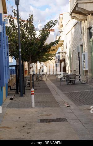 A narrow street lined with buildings primarily constructed of light-colored stone. The street is paved with a mix of light gray and dark gray stones and features a sidewalk on the right side. Several outdoor seating areas with metal tables and chairs are set up along the street, suggesting a cafe or restaurant. A solitary tree stands on the left side of the street, and a street lamp is visible on the left. The sky is a clear blue with a few scattered clouds., Limassol, Cyprus Stock Photo
