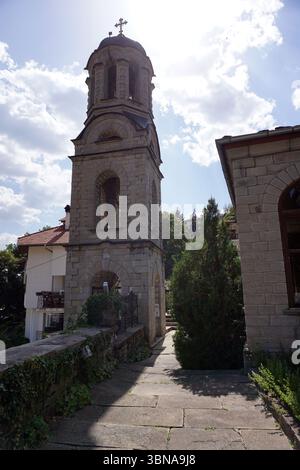Momchilovtsi, a village in the Central Rhodopes in Bulgaria, A serene scene featuring a tall stone bell tower with a cross at the top. The tower, constructed from light-colored stone, stands on a stone path that winds through the area. The tower has a dome-like structure at the top and features arched openings on its sides. A small, dark-colored railing or fence is visible near the base of the tower. To the right of the tower, a stone building with a dark brown or reddish-brown roof is partially visible. The sky is a clear blue with scattered white clouds, and the sun shines brightly, casting Stock Photo