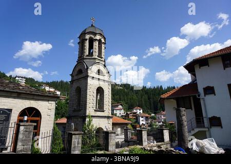 Momchilovtsi, a village in the Central Rhodopes in Bulgaria, A serene church scene. The church, constructed from light-colored stone, stands tall with a bell tower featuring a cross at its peak. The bell tower is adorned with arched windows and a dark gray or black roof. The church is surrounded by a stone wall with a metal fence, and a small, light-colored stone building with a wooden door is visible to the left. The area is nestled amidst a lush green forest, with houses and buildings of varying sizes and colors, including white and red-tiled roofs, scattered throughout the landscape. The Stock Photo