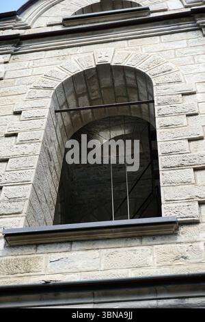 Momchilovtsi, a village in the Central Rhodopes in Bulgaria, A stone archway, likely part of a building, with a window. The window is framed by a dark-colored metal railing or bar. The stone archway is set within a light-colored stone wall, which is made of rectangular blocks. The wall has a slightly uneven texture and a decorative element at the top of the archway. The window is located in the center of the image, and the perspective is from below, looking up at the archway. The sky is visible in the top left corner of the image. An artist's eye & imagination shaped Stock Photo