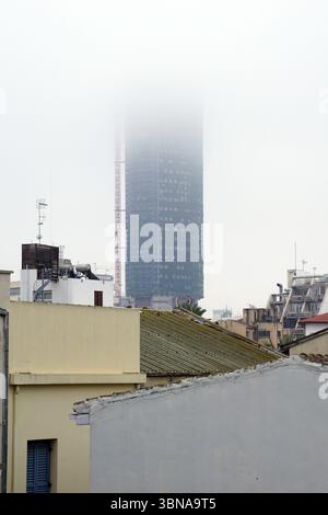 Nicosia (Lefkosia), Cyprus, the world’s last divided capital, A cityscape with a tall, dark blue skyscraper in the background, partially obscured by a thick fog. The skyscraper has numerous windows and appears to be under construction or renovation, with a crane visible on its side. In the foreground, a mix of buildings with various colors and architectural styles, including a yellow building with a flat roof and a white building with a gray roof, are visible. The buildings are partially obscured by the fog, creating a hazy effect. The sky is a light gray or white, and the fog is dense, Stock Photo