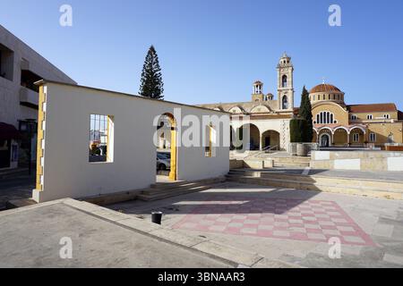 Paralimni, Cyprus, A serene courtyard in front of a church. The courtyard is paved with light-colored stone and features a large, white, rectangular wall with a yellow archway and a small, dark-colored trash can. A checkered pattern of pink and white tiles covers the ground. The church, with its light-colored stone facade and a prominent bell tower, stands majestically in the background. The church has a domed roof and a smaller dome on top. The courtyard is surrounded by a few trees, and a portion of a building with a balcony is visible on the left side of the image. The sky is a clear, Stock Photo