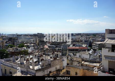 Thessaloniki, Greece. Greece’s second-largest city, located in northern Greece by the Thermaic Gulf. Known for its rich Byzantine, Roman, and Ottoman heritage., The image presents a panoramic view of a densely populated cityscape. The city is characterized by a mix of architectural styles, with buildings exhibiting a variety of colors, including white, beige, and muted tones. Many of the buildings have flat roofs, and some feature chimneys. The cityscape extends towards the horizon, where a range of mountains is visible under a clear blue sky. The vantage point of the image is from a high Stock Photo