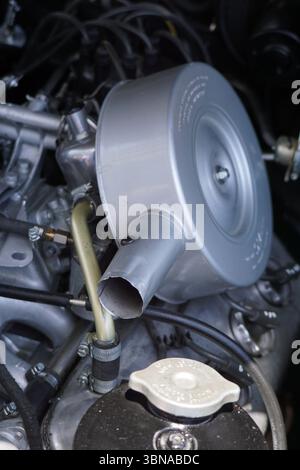 Cyprus, Larnaca, A close-up view of a car engine, with a silver air filter prominently featured in the center. The air filter has a circular opening and a small hole on its side. It is attached to a silver pipe that extends from the engine. The engine is a complex assembly of various metallic components, including hoses, wires, and other parts, all in shades of silver and black. A black cap is visible on the engine, and a small, round, silver cap is also present. The engine is housed within a black plastic cover, and the background is blurred, drawing focus to the engine and its components. Stock Photo