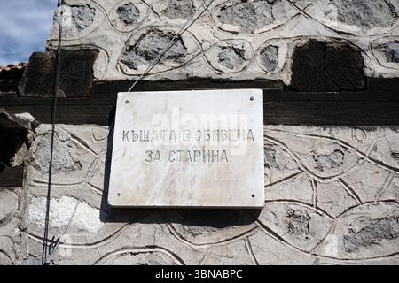 Bansko is a picturesque town in southwestern Bulgaria, nestled at the foot of the Pirin Mountains., A weathered stone wall with a rectangular plaque affixed to it. The plaque is made of a light-colored stone, possibly marble, and features a dark inscription in Cyrillic script. The inscription translates to "The wall is old and the trees are dead." The wall is constructed of irregularly shaped stones, some of which are weathered and worn, and is topped with a dark wooden beam. A wire or cable is visible running along the top of the wall. The background is a clear blue sky. An artist's eye & Stock Photo