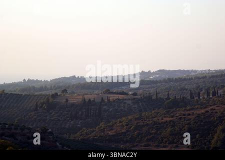 Ağaçlı — a quaint rural neighborhood (mahalle) of Söke district in Aydın Province — lies about 6 km east of Söke centre, nestled in olive‑tree‑studded hills overlooking the Aegean, Turkey, A serene landscape with rolling hills and terraced fields, possibly olive groves, in the foreground. The hills are covered in a mix of green and brown vegetation, with scattered trees and bushes. The sky is a pale, hazy light blue or gray, suggesting either early morning or late afternoon. In the distance, a small settlement is nestled on a hillside, with a few buildings visible. The image is taken from a Stock Photo