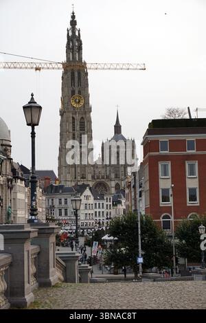 Antwerp, Second-largest Belgian city, located on the Scheldt River in Belgium., A cityscape with a tall, ornate church featuring a clock tower and spires, standing majestically in the background. It is the Cathedral of Our Lady. A construction crane is visible, indicating ongoing development. The foreground is dominated by a cobblestone street, lined with lampposts and trees, and a low stone wall or railing runs along the left side of the street. The buildings in the foreground are primarily brick and white, with some red accents, and feature a mix of architectural styles. A few people can be Stock Photo
