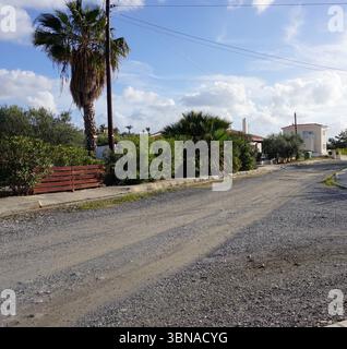 Argaka, Cyprus, A residential street with a gravel road that curves gently to the right. The street is lined with lush green trees and bushes, including a palm tree on the left side. A white house with a red roof is visible in the background, and a white wall or fence runs along the right side of the street. Utility poles and power lines are also visible along the street. The sky is a clear blue with fluffy white clouds. A small, light-colored vehicle is parked on the right side of the Stock Photo