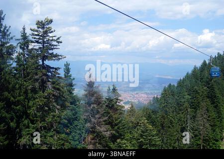 Bansko is a picturesque town in southwestern Bulgaria, nestled at the foot of the Pirin Mountains., A cable car suspended in mid-air, traversing a mountainous landscape. The cable car is primarily blue and white, with a dark-colored railing. It is attached to a cable that stretches across the frame, disappearing into the distance. The cable car is positioned on the right side of the image, moving towards the left. The backdrop features a breathtaking view of a valley, with a settlement nestled below. The valley is surrounded by a dense forest of tall, green trees, and the mountains in the Stock Photo
