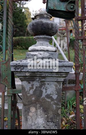 Budapest, Hungary – the enchanting Pearl of the Danube., A weathered stone column with a dark gray/black top and a white band around the middle, standing in a garden setting. The column is part of a larger structure, possibly a gate or fence, with a green metal gate and a section of a dark green/black metal gate visible on the left. The column is surrounded by a mix of green and brown/tan foliage, and a portion of a white structure, possibly a mailbox or post box, is visible in the background. The ground is covered in fallen leaves, suggesting it might be autumn. The background also includes Stock Photo