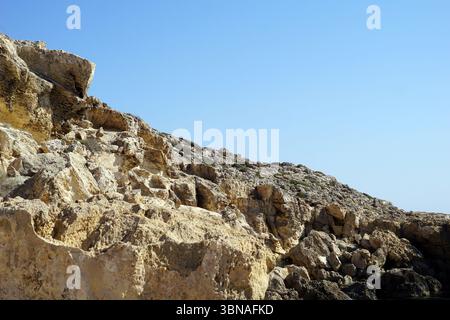Capo Greco, Cyprus, The image depicts a rocky cliff face, primarily composed of light beige and tan rocks, with some darker brown and gray tones. The cliff face is rugged and uneven, with a mix of smooth and rough textures. The perspective is from a low angle, looking up at the cliff, which gives a sense of its height and grandeur. The sky above is a clear, bright blue, providing a stark contrast to the earthy tones of the cliff. The cliff face is bordered by a body of water, which is not visible in the image. An artist's eye & imagination shaped Stock Photo