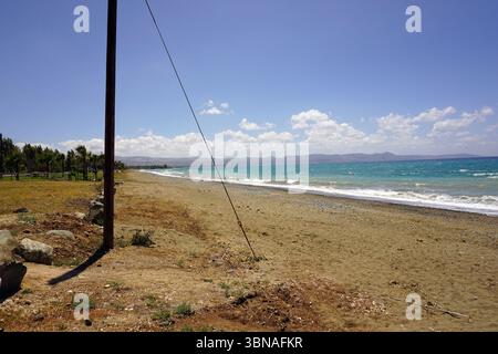 Cyprus Beach, A serene beach scene. The sandy beach stretches out towards the turquoise ocean, where small waves are gently lapping against the shore. The sky is a clear blue, dotted with fluffy white clouds. In the foreground, a utility pole with a dark brown or black pole and a thin wire attached to it stands on the left side of the image. The beach is bordered by a grassy area on the left and a few scattered rocks on the right. In the distance, a line of trees and hills can be seen on the Stock Photo