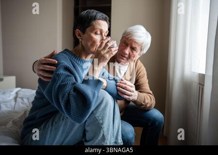 Distressed mature woman in emotional breakdown drinking water, receiving solace from senior partner Stock Photo