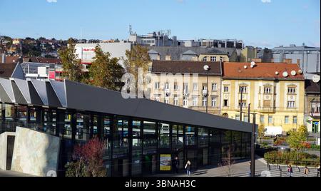 Szell Kalman Ter, Budapest, Hungary – the enchanting Pearl of the Danube., A bustling square filled with people walking and standing, creating a lively atmosphere. In the foreground, a modern building with a distinctive curved roof and large glass windows dominates the scene. The building's facade is primarily a dark gray or black color. The square is surrounded by a variety of buildings, including a yellow building with a red roof and a building with a white facade. The buildings are a mix of architectural styles, with some featuring satellite dishes on their roofs. The square is paved with Stock Photo