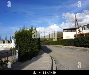 Argaka, Cyprus, A serene residential area with a gravel road curving gently to the right. On the left side of the road, a white house with a red roof is nestled amidst lush greenery, including a neatly trimmed hedge. A small, white, possibly concrete, structure is visible near the hedge. In the distance, a hill covered in vegetation and a few trees can be seen. Power lines stretch across the clear blue sky dotted with fluffy white clouds. A small section of a white wall or structure is visible on the left side of the Stock Photo