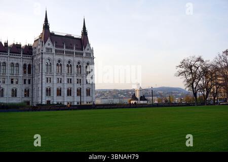 Hungarian Parliament Building. A grand, white building with a red roof and two tall spires, featuring numerous windows and a large, ornate entrance. In the background, a statue of a person stands on a pedestal, and a hill with a mix of residential and possibly commercial buildings is visible. The sky is a light blue with a few clouds, and the scene is bathed in the soft glow of the setting sun, casting a warm light over the entire landscape. A few people can be seen walking on the lawn in the foreground., Budapest, Hungary – the enchanting Pearl of the Danube. Stock Photo