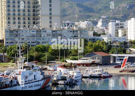A serene harbor scene, with a variety of boats and ships docked in the calm waters. The harbor is nestled amidst a landscape, with a mix of modern and older buildings, including a tall, light-colored building with numerous windows. The buildings are situated on a hillside, with a mountain range visible in the background. The landscape is surrounded by lush greenery, including trees and vegetation. The sky is a clear blue, and the water is relatively calm, reflecting the surrounding buildings and the clear sky. A few cars are parked along the shore, and a small, light-colored building with a re Stock Photo
