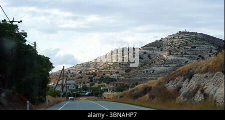 traveling in Cyprus, A winding road cutting through a mountainous landscape. The road, marked by a white line down the center, is flanked by lush green trees and bushes on the left side. On the right side, a steep, rocky hill rises, its surface covered in a mix of light brown and tan colors. A small settlement is nestled at the base of the hill, with houses and buildings visible. The sky above is a light blue, dotted with fluffy white clouds. Power lines stretch across the top of the image, and a car is traveling on the road. An artist's eye & imagination shaped Stock Photo
