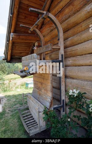 Bulgaria, Gela, A rustic wooden structure, possibly a bench or a small table, attached to the side of a log cabin. The structure is constructed from light brown wooden planks and features a black metal frame. It has a curved top and a flat bottom, with a small wooden shelf on the side. A small, orange-handled tool, possibly a shovel or a small tool, is resting on the shelf. The cabin is made of light brown logs and has a sloping roof. A small section of a green container is visible in the background. The structure is in a grassy area with a few white flowers nearby. An artist's eye & Stock Photo