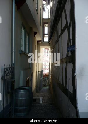 Idstein, Germany, A narrow cobblestone street flanked by buildings with half-timbered facades, characteristic of traditional architecture. The buildings are primarily white or cream-colored with dark brown or black timber framing. A metal gate is visible on the left side of the street, and two dark brown or black wooden barrels are placed on the ground near the gate. A small, dark-colored object, possibly a mailbox, is attached to the wall near the gate. The street is illuminated by natural light filtering through the half-timbered buildings, creating a warm and inviting atmosphere. A small, Stock Photo