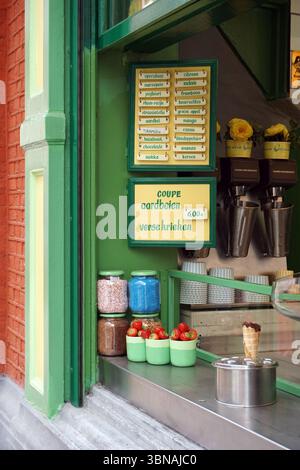 Antwerp, Second-largest Belgian city, located on the Scheldt River in Belgium., A vibrant green storefront with a white sign displaying the text "Coupe aardbeien verse krieken" in Dutch. The storefront is adorned with a variety of colorful jars and containers, including blue, pink, and brown ones, filled with different toppings. A menu board is also visible, listing various ice cream flavors and their prices. A metal ice cream machine is positioned on the right side of the storefront, and a small, round, silver ice cream cone is placed on the counter. A brick building is on the left side, and Stock Photo