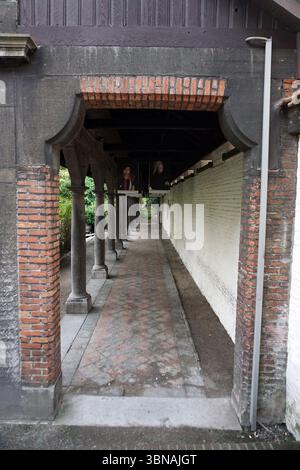 Antwerp, Second-largest Belgian city, located on the Scheldt River in Belgium., A long, narrow walkway with a brick archway at its center. The walkway is lined with brick columns on both sides, creating a sense of depth and perspective. The columns are made of dark gray or black stone and are supported by a dark brown wooden structure. The walkway is paved with a patterned brick or stone surface. On the right side of the walkway, a white wall runs parallel to the path, and a dark-colored, possibly black, statue or sculpture is visible on the wall. The archway is supported by a dark brown Stock Photo