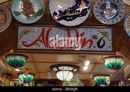 Istanbul, Turkey. The largest city in Turkey and one of the world’s few transcontinental cities, spanning both Europe and Asia across the Bosphorus Strait., A ceiling adorned with a variety of decorative plates and lamps, creating a vibrant and eclectic atmosphere. The ceiling is made of wood and features a grid-like pattern. The plates are predominantly blue and white, with some featuring intricate designs and patterns, including depictions of birds and other figures. The lamps are primarily white with green and blue accents, and some have a stained glass or glass-like appearance. A sign Stock Photo