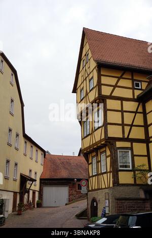 Wertheim,  At the confluence of the Main and Tauber rivers in Baden‑Württemberg, Germany., A quaint town with a cobblestone street lined with buildings in shades of yellow and brown. The buildings feature half-timbered construction, with wooden beams and planks forming a distinctive architectural style. A red-tiled roof adds a pop of color to the scene. A white garage door is visible on the left side of the image, and a small, possibly wooden, structure is located near the garage. A few cars are parked along the street, and a small, possibly wooden, structure is located near the garage. The Stock Photo