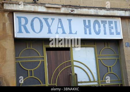 Nicosia (Lefkosia), Cyprus, the world’s last divided capital, The image shows the exterior of a building with a sign that reads "Royal Hotel". The sign is mounted on a beige or tan stone wall. The building's facade features a combination of green and brown elements, including a wooden door with a dark brown or black frame and a white panel with a decorative pattern. The door is partially open, revealing a glimpse of the interior. The building also has a decorative metal frame with a geometric pattern. The building's exterior is made of stone or stucco, and the sign is affixed to the wall with Stock Photo