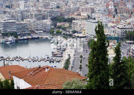Kavala, Greece, The image presents a panoramic view of a cityscape from a high vantage point. The city is densely packed with buildings, predominantly white and gray, with some red-tiled roofs. The cityscape is surrounded by a harbor filled with boats and yachts, and a road runs along the waterfront. The buildings are closely packed together, creating a dense urban environment. The image is taken from a high angle, providing a comprehensive view of the city and its surroundings. The water in the harbor is relatively calm, with a few boats visible. The sky is a light blue, and the trees in the Stock Photo