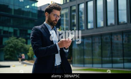 Caucasian businessman man business guy male manager walking in city using mobile phone middle-aged employer entrepreneur working smartphone texting Stock Photo