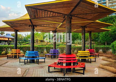 Bright round benches painted in rainbow colors under wooden canopies in a public park. Artistic urban seating surrounded by greenery Stock Photo