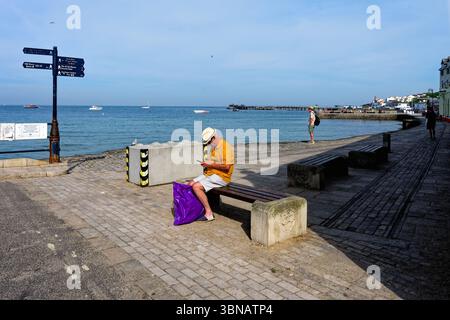 An elderly gentleman sitting on the seafront at Swanage using a mobile phone, with the bay in the background on a sunny summers day Dorset England UK Stock Photo
