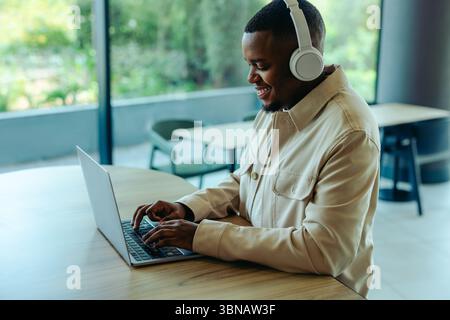 A smiling businessman is working remotely, embodying the digital nomad lifestyle. He wears headphones and types on his laptop, seated in a modern offi Stock Photo
