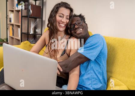 A happy young couple laughs and embraces while using a laptop computer together on the couch at home. Stock Photo