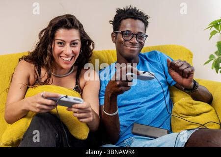 A happy young couple looks at the camera and laughs while playing video games together on their couch. Stock Photo
