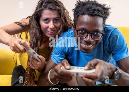 A happy young couple smiles at the camera while playing an energetic video game on the couch at home. Stock Photo