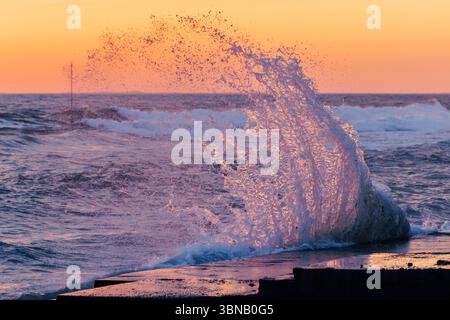 Dramatic sea wave splash at sunset with glowing water droplets and vibrant ocean colors in evening light. Stock Photo