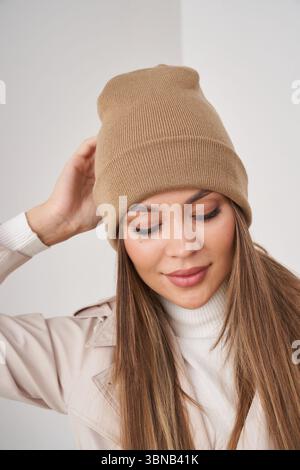 Female wearing a beige knit hat, touching it gently with one hand, dressed in a light coat and white turtleneck, with long hair draping over shoulders Stock Photo