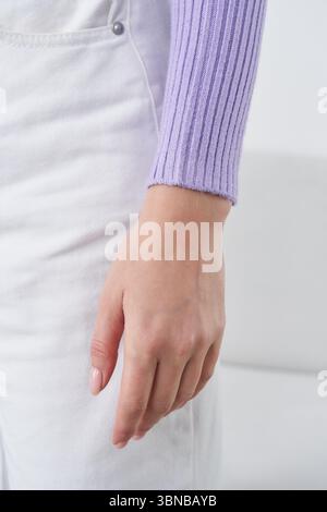 Close-up of a hand with light skin tone, wearing a long-sleeve lilac ribbed sweater, next to light-colored pants with a button detail. The fingers Stock Photo