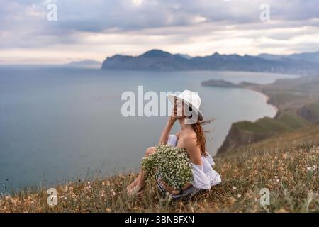 Woman Hat Flowers: Lady with bouquet sits on cliffside at daytime enjoying sea view, resting. Stock Photo